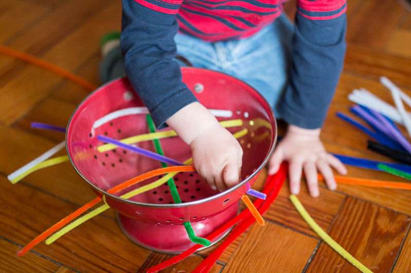 Toddler inserting pipe cleaners in a metal strainer