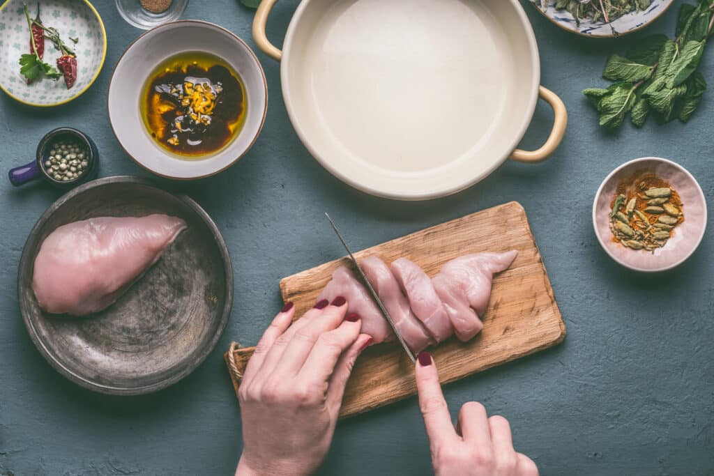 Female hands cut chicken breast on wooden cutting board on kitchen table background with ingredients, top view. Dieting cooking eating and healthy food concept